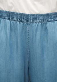 Hele sinised elastse vöökohaga teksapüksid, mida kantakse valge topiga, keskendudes vöökohale ja ülemisele reieosale.