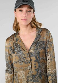 Een bruine blouse met patronen heeft een v-hals, lange mouwen en ingewikkelde paisley-ontwerpen in tinten blauw en wit, gemaakt van een soepele stof.