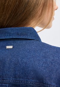 Naise selg helepruuni juustega, seljas sinine teksajakk, mille krae all on väike hõbedane Barbouri logotähis.