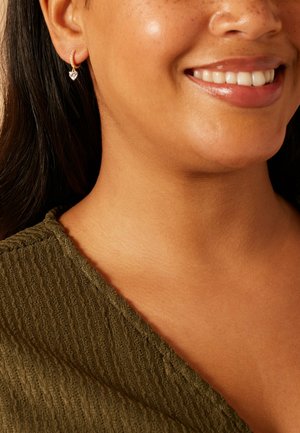 Smiling woman with medium skin tone, dark hair, wearing a green textured top and a small gold hoop earring with a heart-shaped charm.