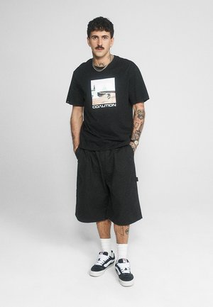 Mężczyzna z tatuażami i wąsami, ubrany w czarną koszulkę Coalition, luźne czarne szorty, białe skarpetki i czarno-białe tenisówki, stojący na jednolitym tle.