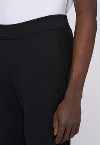 Svarta joggerbyxor med texturerad tyg, elastiskt midjeband, sidofickor och en liten logodetalj på vänster lår.