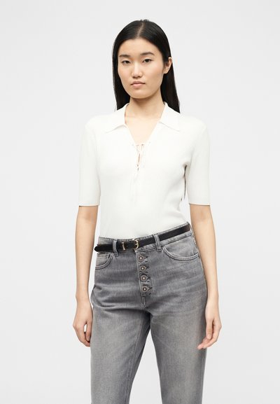 Νεαρή γυναίκα με μακριά μαύρα μαλλιά που φοράει λευκό πλεκτό μπλουζάκι με κοντά μανίκια και γκρι ψηλόμεσο τζιν με διακοσμητικά κουμπιά και μαύρη ζώνη.