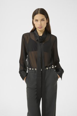 Giovane donna che indossa una blusa nera trasparente con un fiocco a forma di rosa, reggiseno nero, pantaloni grigi e una cintura nera con borchie, con le mani nelle tasche.