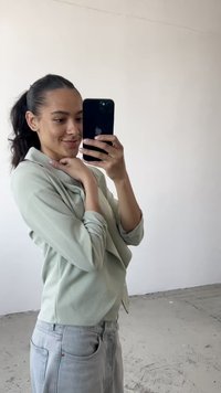 Blusa de manga larga verde claro con cuello drapeado, combinada con jeans gris claro. Tejido suave con un corte holgado. Fondo minimalista.