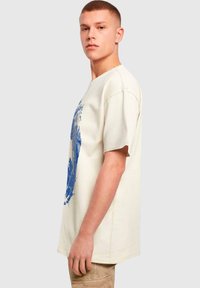 Beige bomull t-skjorte med et blått sprutgrafikk, korte ermer og rund hals, modellert av en mannlig figur i profil.