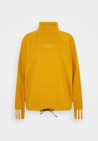 Keltainen fleece-pulloveri, jossa korkea kaulus, puolivetoketju, valkoiset raidat hihoissa ja säädettävä kiristysnauha helmassa.