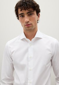 Giovane uomo con capelli ricci e scuri, che indossa una camicia bianca attillata, rivolto verso l'avanti su uno sfondo chiaro e semplice.