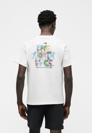 Mies, jolla on lyhyet kiharat hiukset, valkoinen T-paita, jossa on värikäs abstrakti kukkakuvio ja teksti "Never Stop Exploring" takana, yhdistettynä mustiin shortseihin.