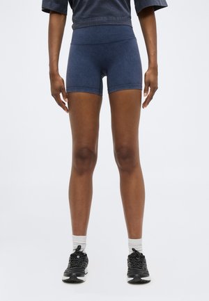 Persoon draagt marineblauwe aansluitende korte broek, zwarte sneakers met witte zolen, witte sokken en een donkergrijze cropped top, staand tegen een effen achtergrond.