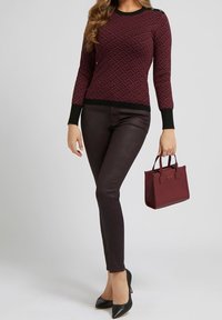 Burgunderfarbener gemusterter Pullover mit schwarzen Akzenten, kombiniert mit dunklen schmalen Hosen und schwarzen spitzen Absätzen. Hält eine kleine burgunderfarbene Handtasche.