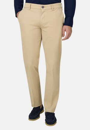 Mand iført beige habitbukser, marineblå loafers og en marineblå skjorte med hænderne delvist i lommerne, stående mod en ensfarvet baggrund.