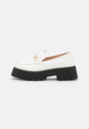 Mocassin en cuir blanc avec semelle épaisse noire à crampons et détail en métal doré sur la tige, présenté en profil sur fond blanc.