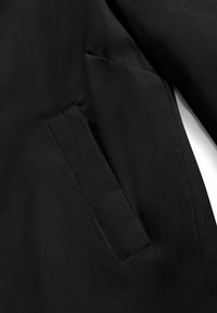 Cappotto nero con una texture liscia e sleek; presenta una tasca laterale e un design minimalista. Il materiale sembra leggero e resistente alle intemperie.