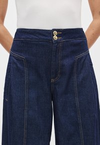 Τζιν παντελόνι με φαρδύ πόδι σε σκούρο μπλε χρώμα και ψηλή μέση, με διπλό κούμπωμα κουμπιών και αντίθετες χρυσές ραφές κατά μήκος των ραφών.