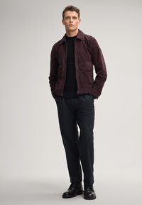 Giacca in suede bordeaux con colletto, due tasche frontali e chiusura con zip, abbinata a pantaloni scuri e stivali in pelle nera.