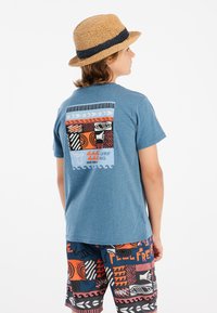Camiseta azul de manga corta con un estampado gráfico colorido en la parte posterior. El niño lleva unos pantalones cortos de baño con estampados y un sombrero de paja.