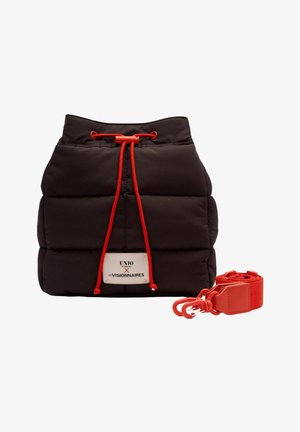 Sac matelassé noir à cordon avec des cordons rouges et une sangle rouge amovible, comportant une étiquette blanche avec du texte.