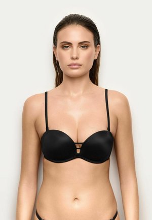 Mujer con el pelo liso y mojado lleva un sujetador negro acolchado con tirantes finos y detalles recortados, de pie frente a un fondo claro y liso.