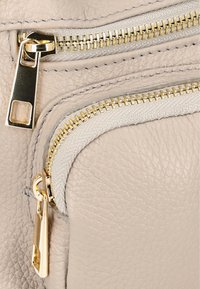 Beige lædertaske med tekstureret overflade, der har guldtonede lynlåse og grå stofdetaljer. Designet inkluderer flere lynlåslommer.