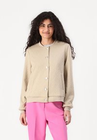 Beige strukturerad jacka med ett fiskbensmönster, rund halsringning och silverfärgade knappar. Har sidofickor samt ribbad fåll och manschetter.