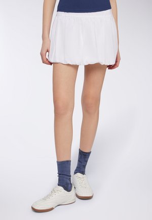 Gonna sportiva bianca a pieghe con forma svasata, abbinata a calze blu a coste e sneakers bianche, mostrata dalla parte superiore della coscia in giù.