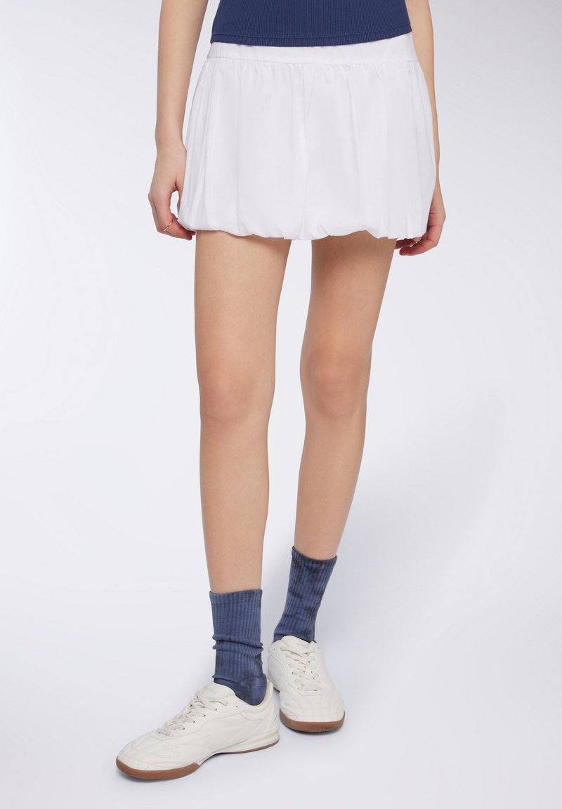 Gonna sportiva bianca a pieghe con forma svasata, abbinata a calze blu a coste e sneakers bianche, mostrata dalla parte superiore della coscia in giù.
