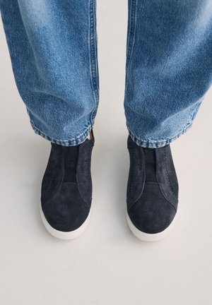 Fekete velúr slip-on cipőt viselő lábak fehér talppal, világos padló háttér előtt, kék farmernadrággal párosítva.