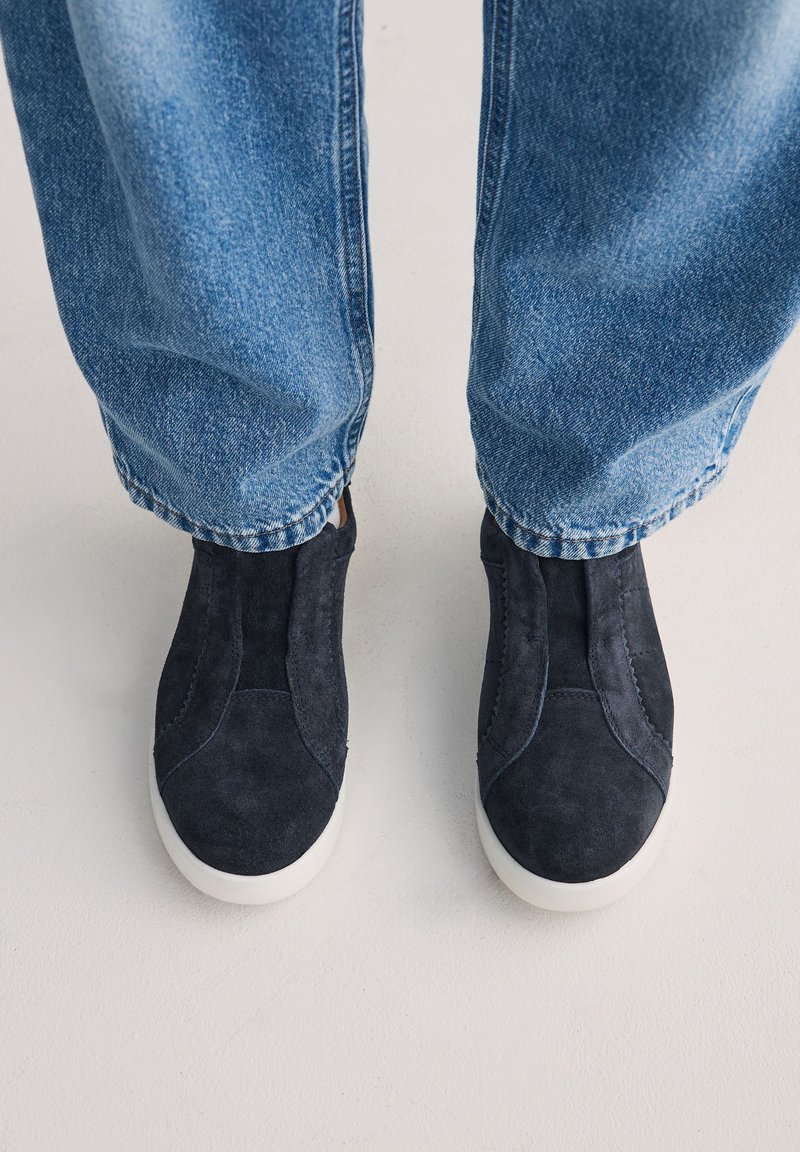Pieds portant des chaussures slip-on en suède noir avec semelles blanches, assorties à un jean bleu sur un fond de sol clair.