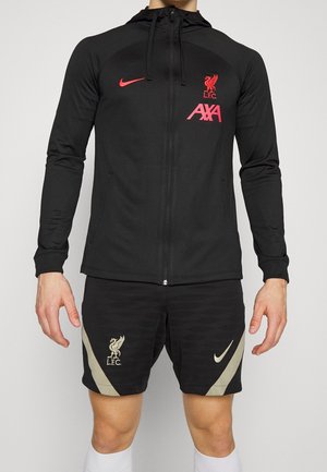 Osoba ubrana w czarną sportową kurtkę Liverpool FC z czerwonymi logo oraz czarne spodenki z beżowymi akcentami i logo Liverpool oraz Nike.