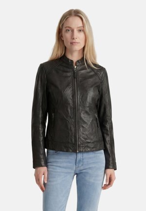 Frau mit langen blonden Haaren, die eine schwarze Lederjacke mit Reißverschluss und hellblaue Jeans trägt, steht vor einem schlichten hellen Hintergrund.