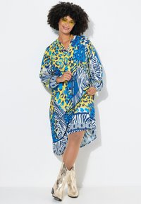 Kleurrijke jurk met blauwe, gele en witte dierenpatronen. Heeft een ontspannen pasvorm, lange mouwen en een strikdetail in de taille.