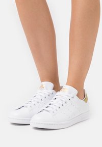 Vita Adidas Stan Smith-sneakers med guldaccenter, barfota mot en enfärgad vit bakgrund.
