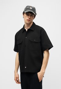 Fiatal férfi fekete rövid ujjú, mellzsebes gombos ingben és fekete baseballsapkában, egy egyszínű szürke háttér előtt áll.