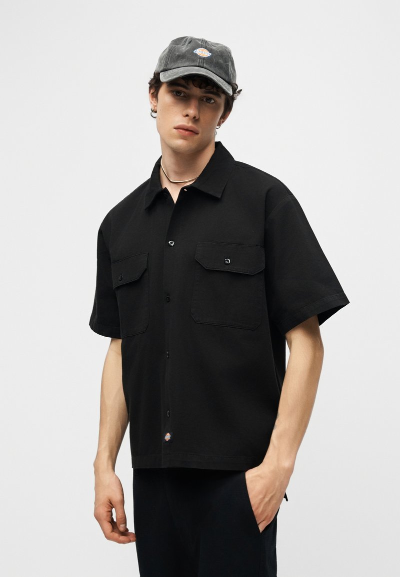 Giovane uomo con una camicia nera a maniche corte con tasche sul petto e un cappellino da baseball nero, in piedi davanti a uno sfondo grigio uniforme.