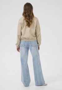 Veste bomber beige à manches longues, avec des poignets et un ourlet côtelés, associée à un jean bleu clair à taille haute et à jambe large avec des poches arrière.