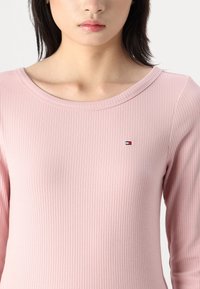 Różowa, prążkowana bluzka z długim rękawem, o szerokim dekolcie i małym, haftowanym logo w czerwonym, białym i niebieskim kolorze na piersi.