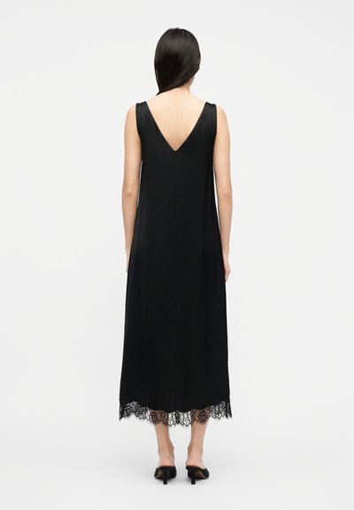 Γυναίκα που φοράει μαύρο midi φόρεμα χωρίς μανίκια με V-πλάτη και δαντέλα στο τελείωμα, σε συνδυασμό με μαύρα mules με τακούνι, στέκεται μπροστά σε απλό φόντο.