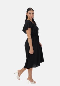 Vestido negro con mangas cortas con volantes, cintura ceñida y falda larga hasta la rodilla. La modelo lleva sandalias de tacón blanco y elegantes.