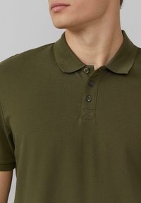 Oliwkowa koszulka polo z teksturowanej tkaniny, z klasycznym kołnierzykiem i zapięciem na trzy guziki z ciemnymi guzikami oraz wyszytym detalem przy szyi.