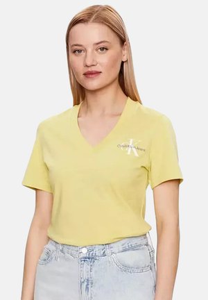 Femme portant un t-shirt jaune à col en V Calvin Klein et un jean clair, debout sur un fond uni.