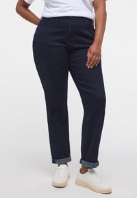 Tumši džinsi ar taisnu kāju, ar sarullētām apmalēm un jostas cilpām, apvienoti ar baltām sporta apaviem ikdienišķam izskatam.