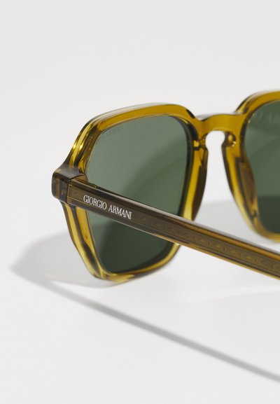 Ochelari de soare pătrați cu rame transparente de culoare verde măsliniu, lente verzi închise și temple texturate de culoare maro, inscripționate cu "Giorgio Armani".