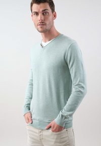 Pull en maille de couleur bleu canard clair avec un col en V et trois boutons décoratifs. Équipé de manches longues et de poignets et d'ourlets côtelés. Porté par-dessus un t-shirt blanc.