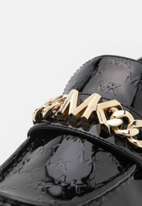 Mocassin en cuir verni noir avec motif de logo en relief, présentant un détail de chaîne dorée et de grosses lettres dorées sur la sangle.