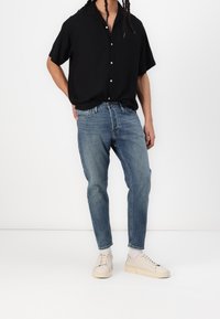 Osoba w czarnej koszuli na krótki rękaw z guzikami, niebieskich dżinsach, białych skarpetkach i beżowych trampkach, stojąca z jedną ręką za plecami.