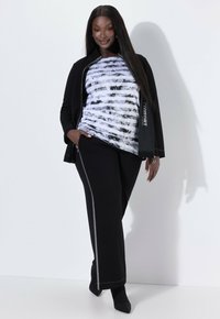 Kobieta ubrana w czarną kurtkę, czarne spodnie z srebrnym paskiem, czarne buty i biało-czarny pasiasty top, stojąca na tle białej ściany.