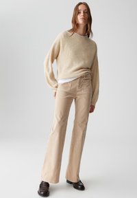 Giovane donna che indossa un maglione beige, pantaloni chiari a campana e mocassini marroni scuri, in piedi contro uno sfondo chiaro e neutro.