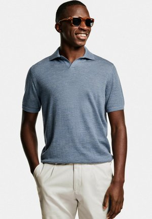 Lachende man met bruine zonnebril, een blauw poloshirt met korte mouwen en lichtbeige broek, handen in de zakken tegen een effen achtergrond.