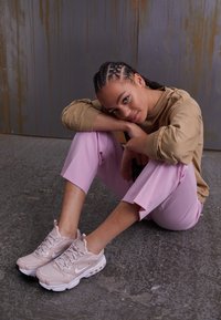 Scarpe da ginnastica rosa chiaro con mesh testurizzato e dettagli in gomma, abbinate a pantaloni larghi rosa pastello e una felpa beige. Sfondo grigio.